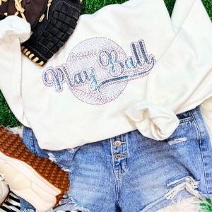 May include: A white sweatshirt with the words "Play Ball" in a rhinestone design, set against a baseball graphic. The sweatshirt is paired with denim shorts and sneakers, with a baseball glove in the background. The overall theme is baseball.