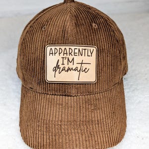 May include: Brown corduroy baseball cap with a rectangular patch. The patch reads "Apparently I'm dramatic" in black script and block letters. The cap has a curved brim and a structured crown.