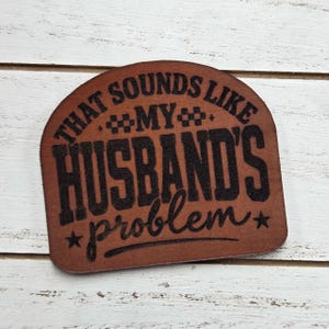 May include: A brown, semi-circular wooden plaque with the phrase "THAT SOUNDS LIKE MY HUSBAND'S problem" in black lettering. The plaque has a rustic, engraved appearance, with stars and decorative elements.