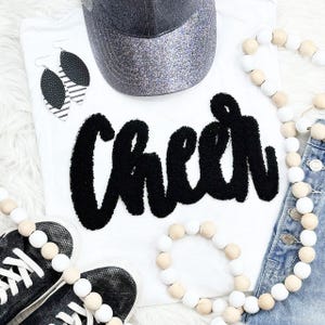 May include: A white t-shirt with the word "Cheer" in large, black, textured letters. The image also includes a silver glitter baseball cap, black and white earrings, black sneakers, and beaded necklaces.
