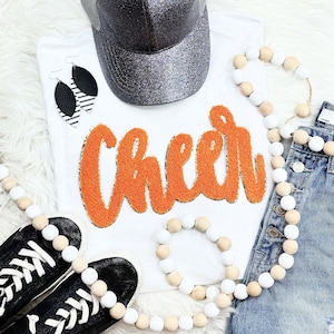 May include: A flat lay featuring a white t-shirt with the word "Cheer" in orange, a glittery gray baseball cap, black sneakers, and denim shorts. Accessories include black and white earrings and wooden bead garlands.