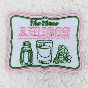 May include: Embroidered patch with a white background, featuring the text "The Three AMIGOS" in pink. Below the text are green embroidered images of a salt shaker, a shot glass, and a lime wedge. The patch has a pink and green border.