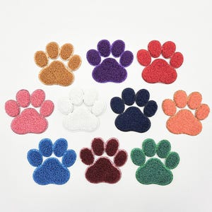May include: A collection of colorful paw print patches in various colors, including brown, purple, red, pink, white, navy, orange, blue, maroon, and green. These patches are likely for pet-themed crafts or apparel.