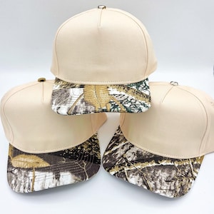 May include: Three beige baseball caps with camouflage print brims are stacked and arranged. The caps have a classic six-panel design with a curved brim. The camouflage pattern features shades of brown, green, and white.