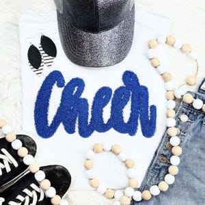 May include: White t-shirt with the word "Cheer" in blue textured lettering. The image also includes a black glitter baseball cap, black and white earrings, black sneakers, and beaded garlands. Denim shorts are also visible.