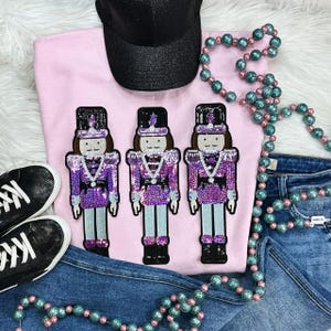 May include: A light pink sweatshirt with three sequined nutcracker figures in purple, black, and brown. The image includes a black glitter baseball cap, blue jeans, black sneakers, and beaded necklaces.
