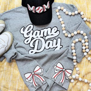 May include: A gray sweatshirt with the words "Game Day" in white script. The sleeves feature baseball bow designs. A black baseball cap with "MOM" and a baseball design is above the sweatshirt. White sneakers and a wooden bead garland complete the look.