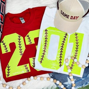 May include: Red and white softball-themed t-shirts with large neon yellow numbers and baseball stitching details. A tan baseball cap with "GAME DAY" text is also visible. A string of wooden beads adds a decorative touch.