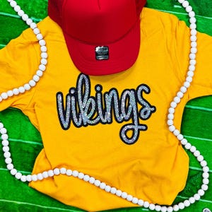 May include: A yellow t-shirt with the word "Vikings" in silver sequins, a red trucker hat, and a white beaded necklace. The shirt is laid on a green surface.