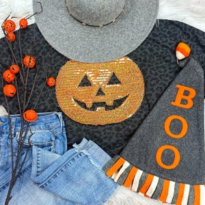 May include: A Halloween-themed ensemble featuring a gray hat, a black top with a sequined pumpkin design, and a gray "BOO" hat. Light blue jeans and decorative orange pumpkins complete the festive look. The hat has a gold band.