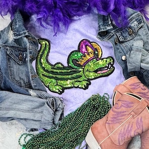 May include: A light purple t-shirt with a sequined green alligator wearing a crown, paired with a denim jacket, pink cowboy boots, green beads, and a purple feather boa. The image evokes a festive theme.