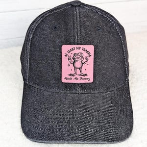 May include: A black denim baseball cap with a pink square patch. The patch features a cartoon frog giving peace signs, with the text "At Least My Trauma Made Me Funny."