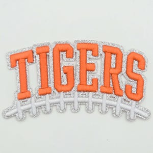 May include: An embroidered patch featuring the word TIGERS in bold orange block letters with a silver glitter border. Below the text is a white embroidered graphic of a football lace design, also outlined in silver glitter.