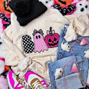 May include: A cream-colored sweatshirt with a Halloween-themed design featuring the word "BOO" with a checkered "B", a pink ghost, and a pink pumpkin. The image also includes a black pom-pom beanie, blue jeans, and sneakers with pink accents.