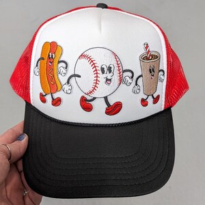 May include: A baseball-themed trucker hat with a white front panel featuring embroidered cartoon images of a hot dog, baseball, and coffee. The hat has a black brim, red mesh sides, and a black rope detail.
