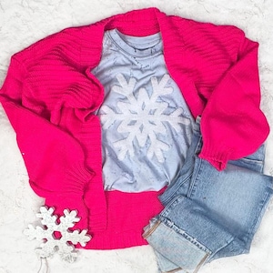 May include: A bright pink, chunky-knit cardigan draped over a light blue t-shirt with a large white snowflake design. Light wash denim jeans are folded next to the sweater and shirt. A silver glitter snowflake decoration is in the foreground.