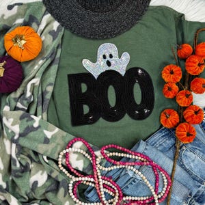 May include: An olive green sweatshirt with a sequined "BOO" design and a sparkly ghost. The image includes a black hat, camouflage print blanket, denim jeans, orange and purple pumpkins, and a pink and white beaded necklace. A fall-themed decorative arrangement.