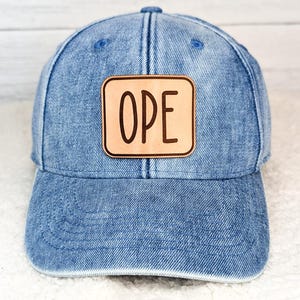 May include: A blue denim baseball cap with a curved brim. The cap features a light brown leather patch with the word "OPE" in dark brown letters. The cap has a classic six-panel design and an adjustable strap.