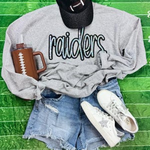 May include: A gray sweatshirt with the word "raiders" in iridescent lettering, paired with denim shorts, white sneakers, and a football-themed mug and cap. The cap is black with a football design.