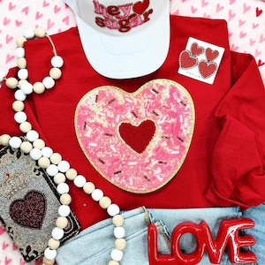 May include: A red long-sleeve shirt with a pink heart-shaped sequined patch, a white trucker hat with "hey sugar" text, and a pair of heart-shaped earrings. A red "LOVE" sign and a beaded garland are also present.