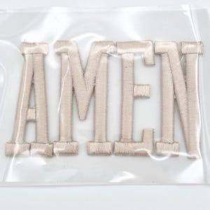 May include: A set of beige embroidered letters spelling the word AMEN, featuring a block font style with a satin stitch finish. The letters are enclosed within a clear, protective plastic packaging sleeve, showing a smooth, uniform texture.