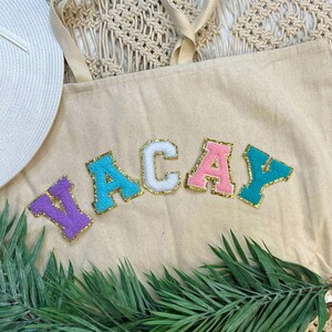 May include: A beige canvas tote bag with the word "VACAY" appliqued in colorful, textured letters. The letters are outlined in gold glitter. A white sun hat and green palm leaves are visible.