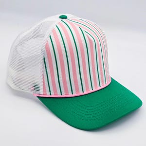 May include: A trucker hat with a green brim and a white mesh back. The front panel features vertical stripes in pink, green, and white. A pink cord accents the brim, and a green button tops the hat.