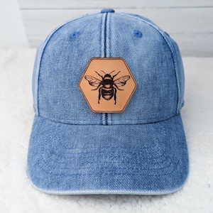 May include: Blue denim baseball cap with a brown leather hexagon patch featuring a detailed bee illustration. The cap has a curved brim and adjustable strap, offering a casual and stylish accessory.