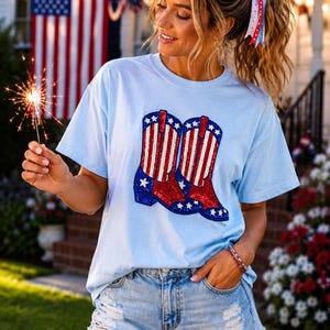 May include: A light blue short-sleeve t-shirt featuring a graphic of two cowboy boots decorated with red and white vertical stripes and blue sections with white stars. The graphic is rendered in a sequined texture, creating a patriotic design.
