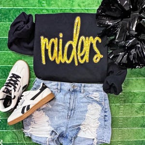 May include: Black sweatshirt with "raiders" in gold glitter lettering, paired with distressed denim shorts, white sneakers with black accents, and a black pom-pom. The items are arranged on a green football field background.