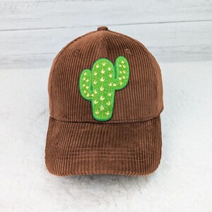 May include: A brown corduroy baseball cap featuring a green cactus patch. The cactus is embroidered with white details and outlined in green. The cap has a curved brim and a structured crown.