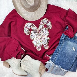 May include: A burgundy sweatshirt with a sequined candy cane and bow design. The image also includes a tan cowboy hat, blue jeans, and cream-colored ankle boots. The outfit is arranged on a white, fluffy surface.