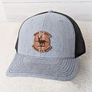 May include: Gray trucker hat with a black mesh back. The hat features a brown leather patch with a deer silhouette inside a crosshair design. The patch has the words "BORN TO HUNT" and "FORCED TO WORK" in a circular arrangement.