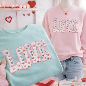 May include: A light blue sweatshirt and a pink sweatshirt, both featuring the word "LOVE" embroidered with colorful heart-shaped accents. The sweatshirts are displayed on a mannequin and held by a person.