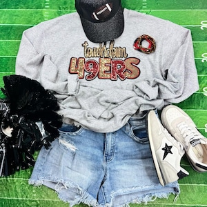 May include: A gray sweatshirt with the text "Touchdown 49ers" in gold and red glitter, paired with denim shorts, sneakers, a black baseball cap with a football design, and a black pom-pom. The items are arranged on a green background.