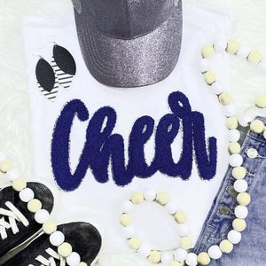 May include: White t-shirt with the word "Cheer" in navy blue, paired with a glittery gray baseball cap, black and white earrings, black sneakers, and beaded jewelry. The ensemble suggests a cheerleading or spirit theme.