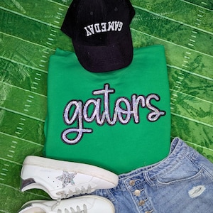 May include: A green sweatshirt with the word "gators" in silver sequins, a black corduroy "GAMEDAY" baseball cap, white sneakers with silver accents, and denim shorts. The items are arranged on a green background with white lines.