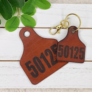 May include: Two brown leather tags with the number "50125" printed in black. One tag is a keychain with a gold-colored ring and clasp. The tags are on a white wooden surface with green leaves in the background.