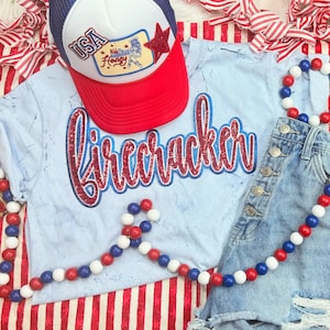 May include: A patriotic ensemble featuring a light blue t-shirt with the word "Firecracker" in red glitter, paired with denim shorts and a red, white, and blue trucker hat. Decorative wooden beads in red, white, and blue complete the look.