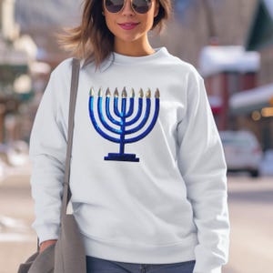 May include: White sweatshirt featuring a blue sequined menorah design. The menorah has eight candle holders with gold accents. The sweatshirt is a crew neck style and is worn by a person.