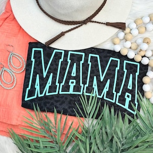 May include: Black t-shirt with the word "MAMA" in large, light blue block letters. The shirt has a leopard print design. The image also includes a white hat with a brown band, orange shorts, and turquoise earrings.