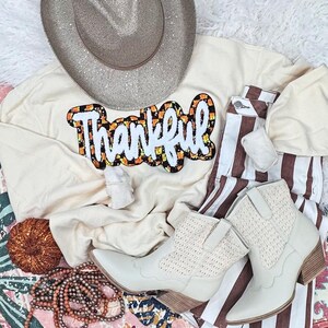 May include: An ivory-colored t-shirt with the word "Thankful" in a candy corn-themed design, paired with brown and white striped pants, cream-colored cowboy boots, and a silver-toned cowboy hat. A fall-themed ensemble.