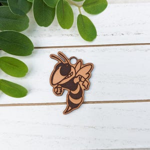 May include: A wooden charm in the shape of a cartoon bee, featuring brown and tan colors. The bee has a detailed design with wings, antennae, and a stinger. The charm has a small hole at the top for hanging.