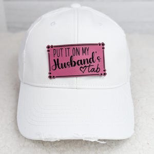 May include: White baseball cap with a distressed brim. A pink rectangular patch on the front reads "PUT IT ON MY Husband's Tab" in black script and block letters, with a heart symbol.