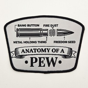 May include: A light gray embroidered patch with a black border featuring a diagram of a bullet. The text reads BANG BUTTON, FIRE DUST, METAL HOLDING THING, FREEDOM SEED, and ANATOMY OF A PEW. The design is centered on a shield-shaped fabric base.