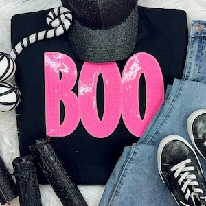 May include: Black sweatshirt with the word "BOO" in bright pink sequins. The image also includes a black glitter baseball cap, light wash denim jeans, and black sneakers with white laces.
