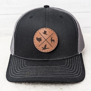 May include: Black and gray trucker hat with a brown leather patch. The patch features a turkey, a bird, a deer, and a duck, with a cross design. The hat has a curved brim and a mesh back.