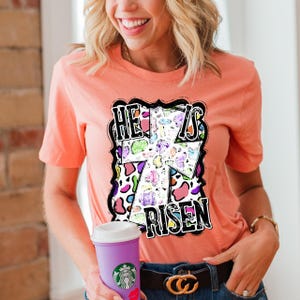 May include: Peach-colored t-shirt with the text "HE IS RISEN" in a black, white, and colorful design. The design features a cross shape with a mix of patterns and colors. The person is holding a purple cup.