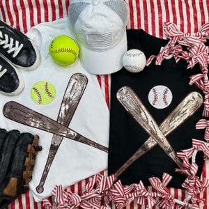 May include: A baseball-themed arrangement featuring a white t-shirt with a sequined baseball bat design, a black t-shirt with a baseball and bat design, a white baseball cap, a softball, a baseball, and a baseball glove. The background has red and white stripes.