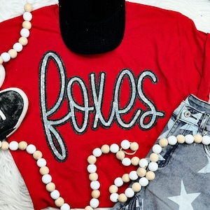 May include: A red t-shirt with the word "Foxes" in silver sequins. The shirt is paired with star-patterned denim jeans, a black cap, and a beaded necklace. A black and white sneaker is also visible.